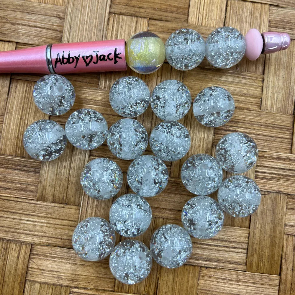 16MM silver glitter crackle acrylic beads with crushed foil effect displayed on woven surface next to a beadable pen, showing approximately 20 clear silver sparkle beads.