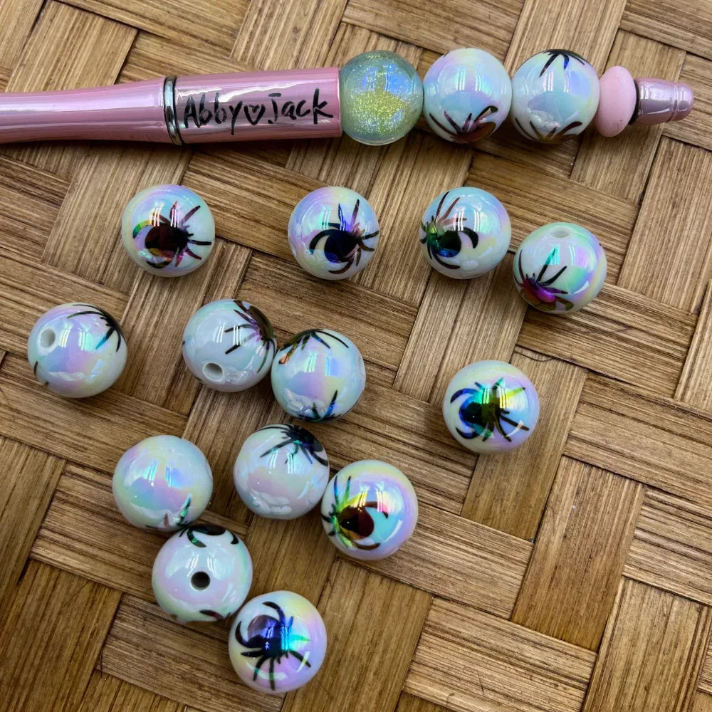 16MM white iridescent AB acrylic beads with colorful spider print design, shown loose on wicker surface and threaded on a pink beadable pen labeled Abby Jack