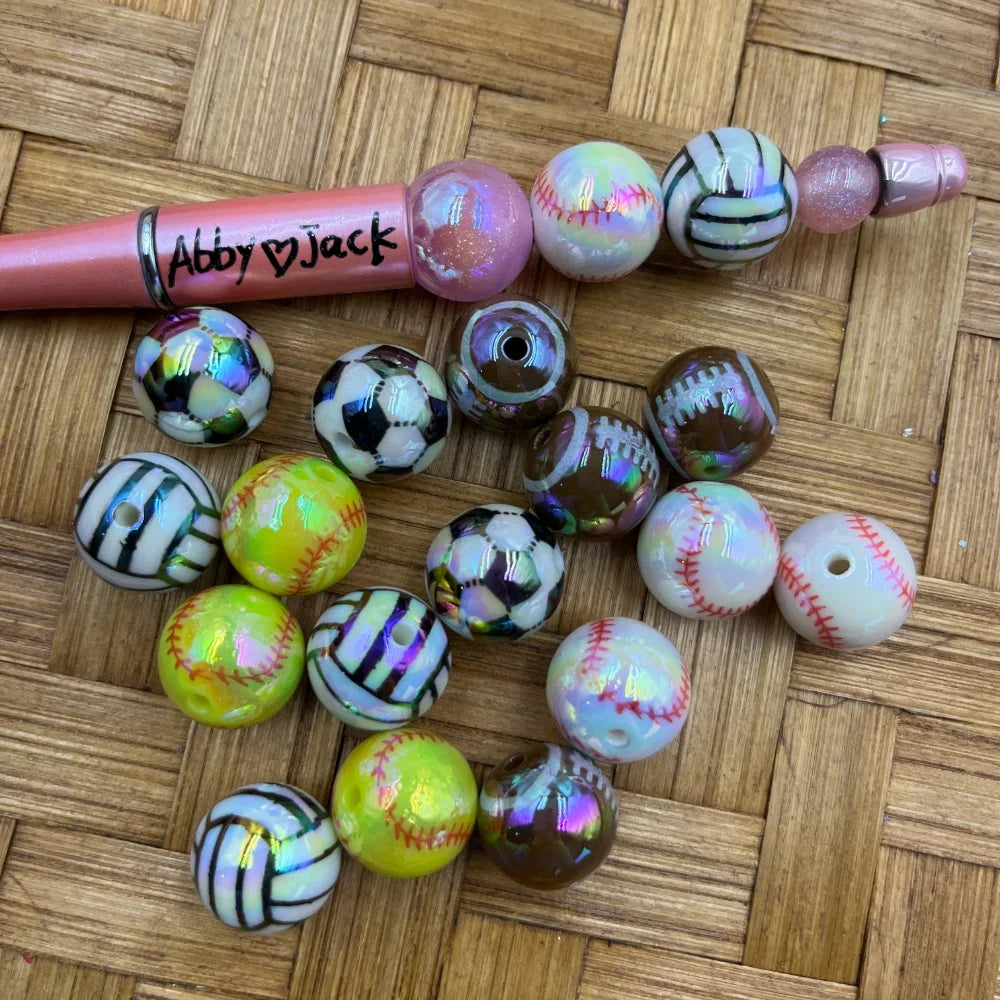 16MM iridescent AB sports acrylic beads featuring baseball, softball, soccer ball, volleyball, and football designs in aurora borealis finish, shown with a beadable pen on a wicker surface.