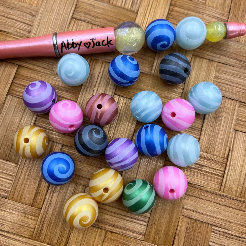 16MM multicolor swirl stripe acrylic beads in assorted colors including pink, blue, purple, gold, and gray with spiral pattern design, displayed on wicker surface next to a beadable pen.