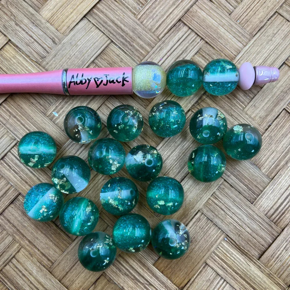 16MM teal transparent acrylic beads with gold flake glitter inclusions displayed on woven surface next to a pink beadable pen labeled Abby and Jack