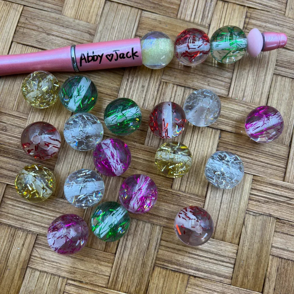 16MM clear acrylic round beads filled with colorful tinsel confetti strands in pink, green, red, yellow, and clear, displayed on a woven surface next to a pink beadable pen labeled Abby Jack.