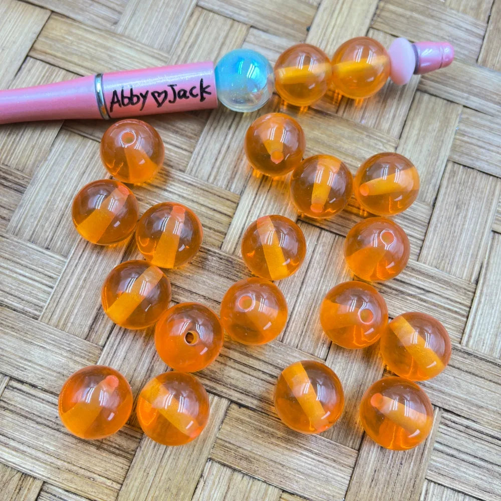 16MM transparent orange round acrylic beads with glossy finish displayed on a woven surface next to a pink beadable pen, showing drilled holes for DIY jewelry making.