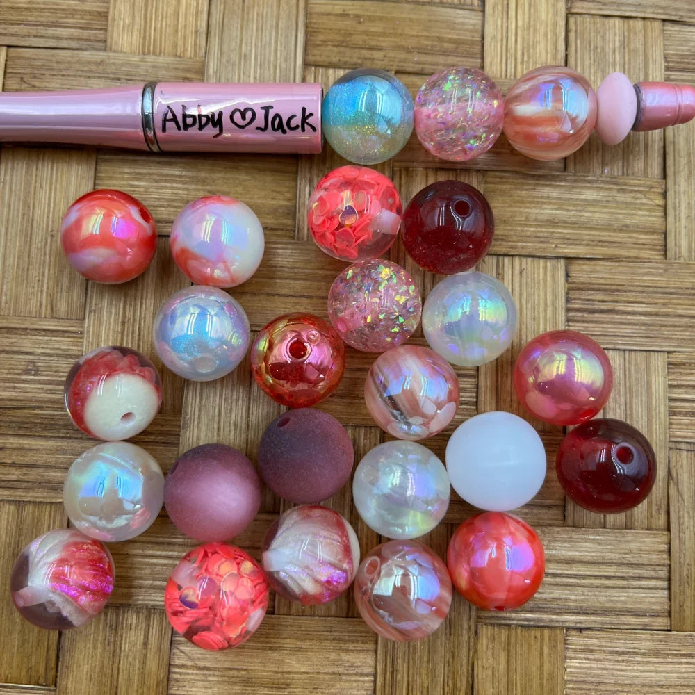 16MM Valentine pink and red iridescent acrylic beads in assorted finishes including AB glitter, marble, confetti, matte, and translucent styles displayed next to a pink beadable pen on a wicker surface.