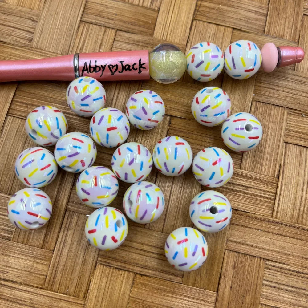 16MM white acrylic beads with colorful rainbow sprinkle donut pattern, shown with a pink beadable pen on a wicker surface from DIY Mini Planet.