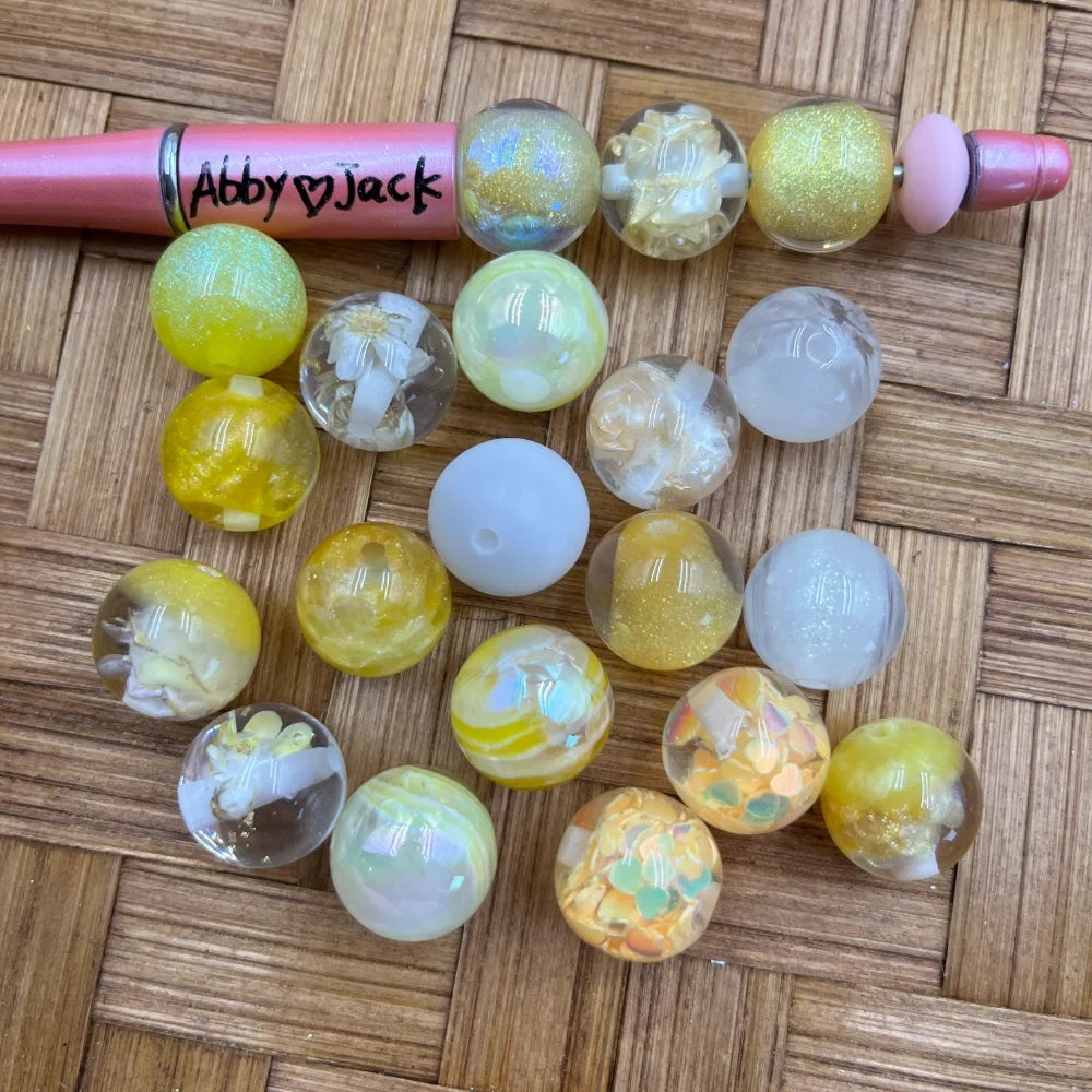 16MM yellow and white acrylic beads in assorted styles including glitter, iridescent, dried floral, confetti, and solid finishes displayed on a woven surface next to a pink beadable pen labeled Abby Jack.