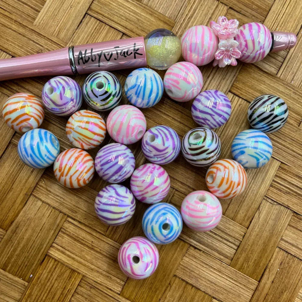 16MM iridescent zebra stripe acrylic beads in assorted colors including pink, purple, orange, blue, and black on white base, displayed next to a pink beadable pen on a woven surface.