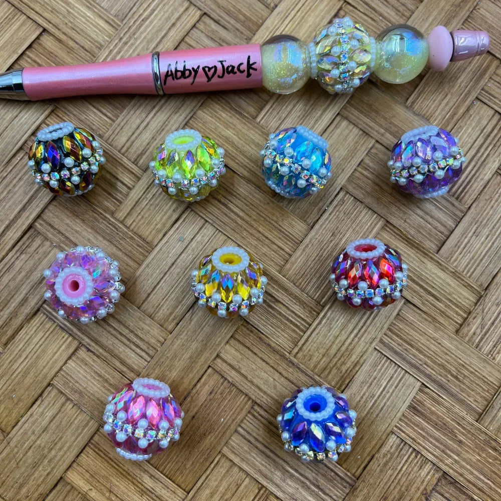 Colorful iridescent AB crystal rhinestone and pearl fancy beads in pink, blue, purple, yellow, and red displayed on a woven surface next to a decorated beadable pen labeled Abby Jack.
