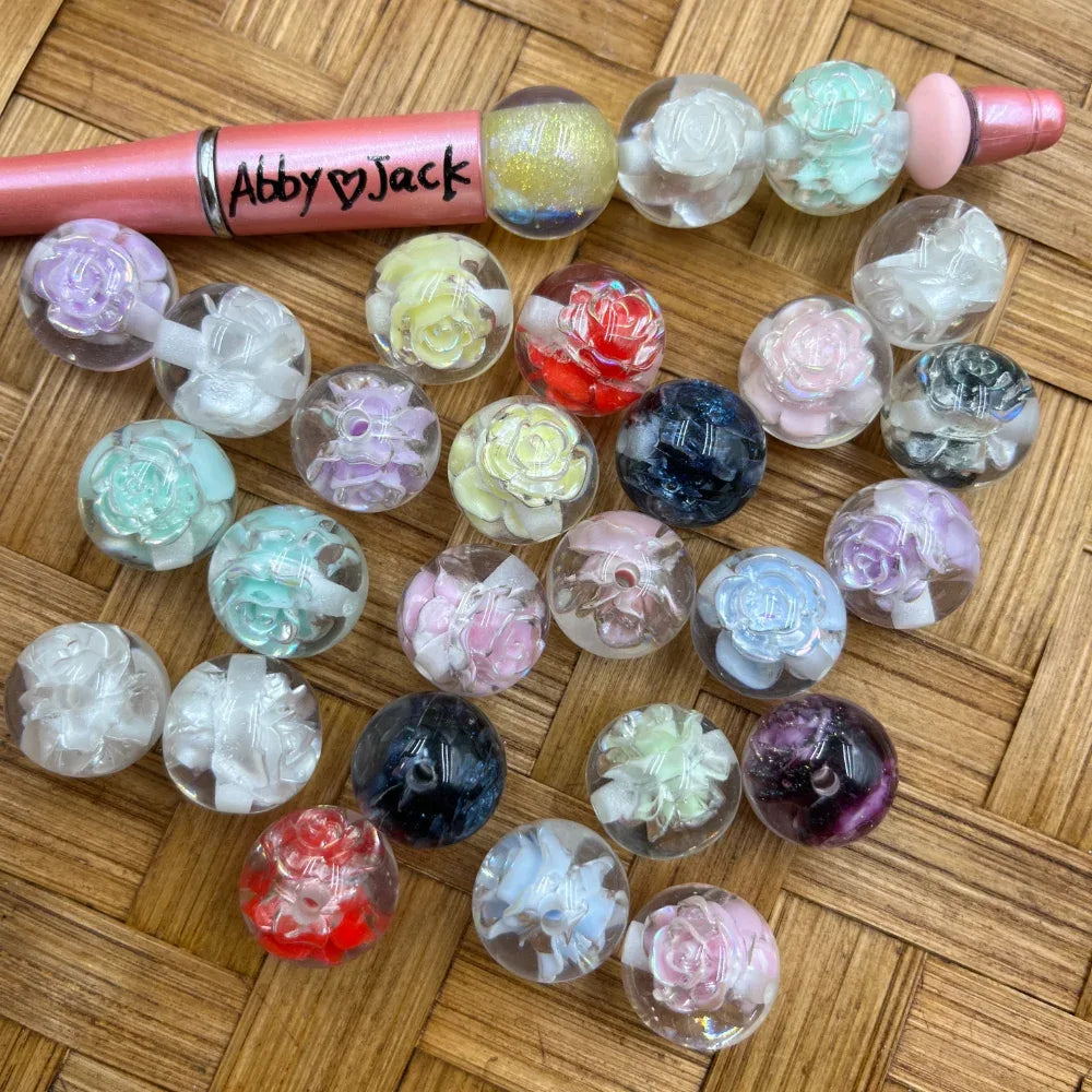 20MM clear acrylic round beads with encased 3D rose flowers in assorted colors including white, pink, mint, navy, red, purple, and black displayed on wicker surface next to a beadable pen.