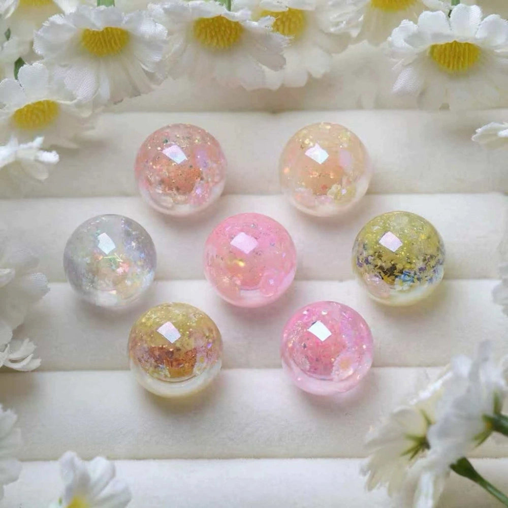 Six round iridescent glitter galaxy acrylic beads in pastel pink, yellow, and clear with holographic sparkle inclusions, arranged on a white surface surrounded by daisy flowers.