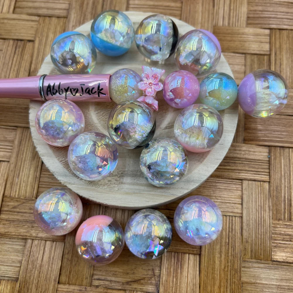 20MM aurora iridescent holographic crystal resin sphere pen toppers in pastel pink, blue, purple, and clear with sparkling faceted crystal inclusions, displayed on a wooden tray with a pink beadable pen.