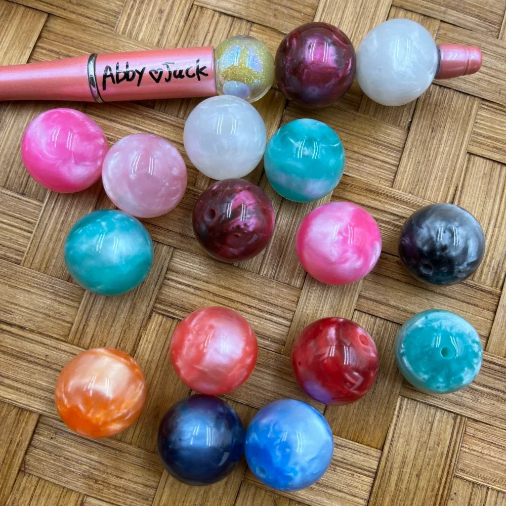 20MM bowling ball shaped acrylic beads in assorted marbled swirl colors including pink, teal, red, orange, navy, white, and galaxy finishes displayed on a woven surface next to a beadable pen.