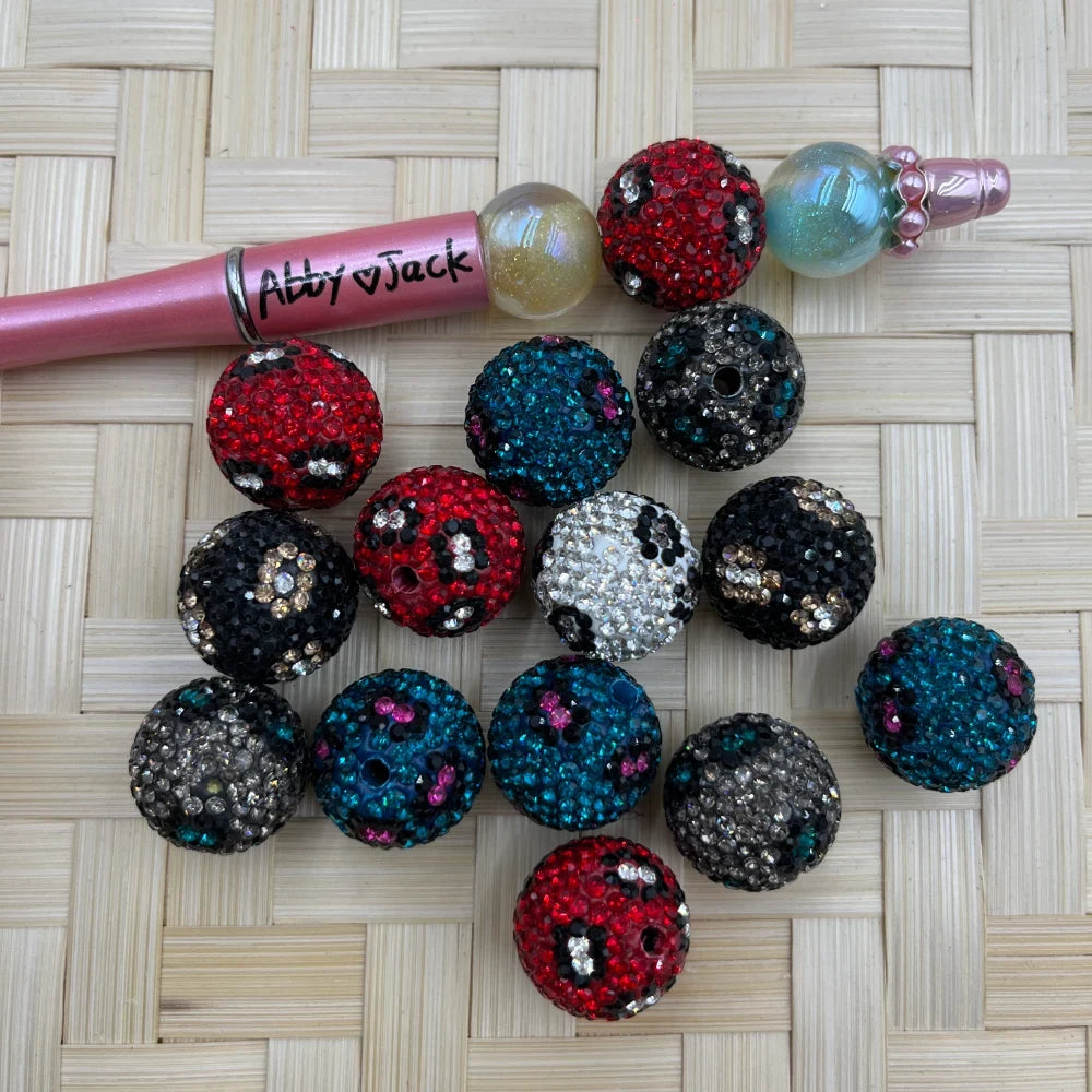 20MM leopard print clay rhinestone beads in red, teal, black, and silver colorways displayed on a woven surface next to a beadable pen.