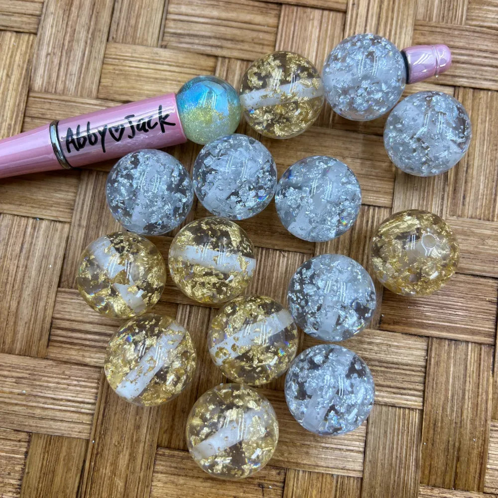 20MM clear acrylic round beads with gold and silver foil flakes, displayed next to a pink beadable pen on a wicker surface, showing metallic flake inclusions in transparent resin-style finish.