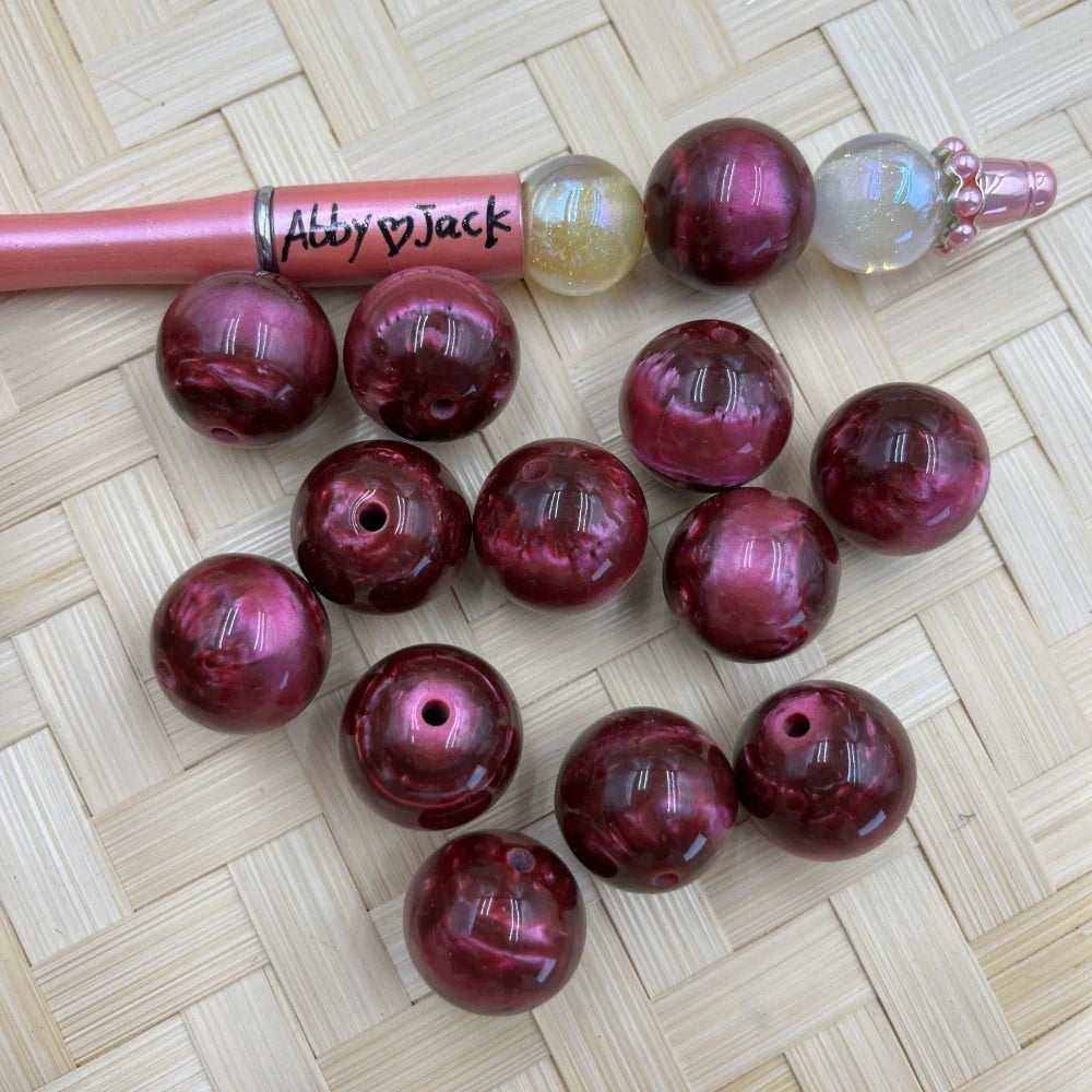 20MM dark crimson burgundy pearl swirl acrylic beads with glossy finish displayed on woven surface next to a beadable pen, showing drilled holes for DIY jewelry making.