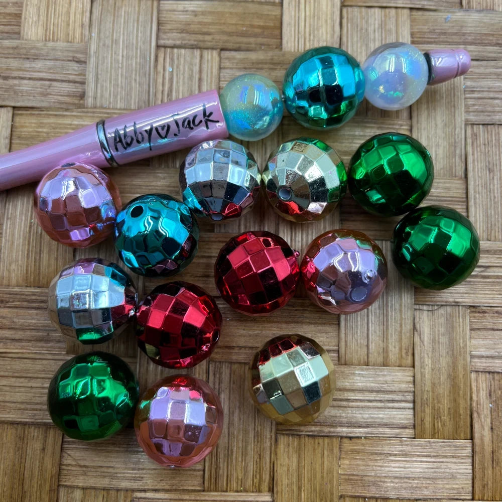 20MM disco ball mirror acrylic beads in red, green, silver, teal, and gold metallic faceted finishes displayed on woven bamboo surface next to a pink beadable pen.