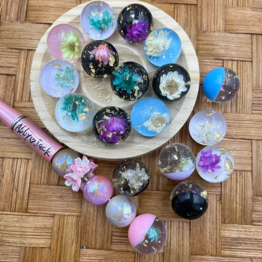 20MM dried flower and gold foil resin sphere pen toppers in black, blue, purple, and clear colors displayed on a wooden tray alongside a beadable pen, craft beads on a wicker surface.
