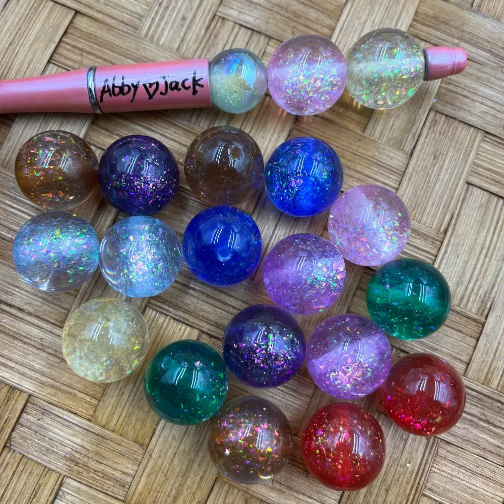 20MM round acrylic beads filled with iridescent galaxy glitter in assorted colors including blue, purple, green, red, amber, clear, and pink, displayed on a woven surface next to a pink beadable pen.