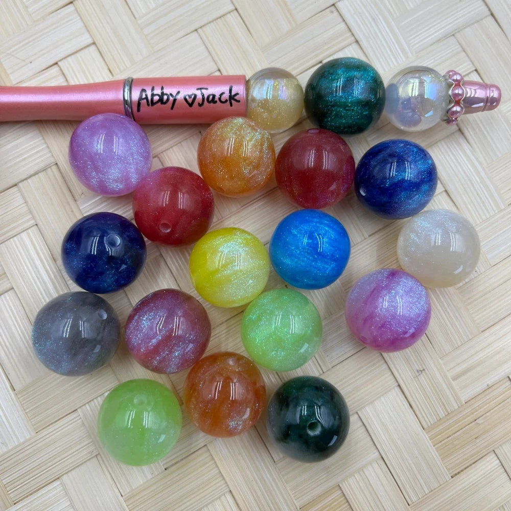20MM galaxy glitter acrylic beads in assorted colors including teal, blue, pink, red, yellow, green, orange, and purple with shimmer finish, displayed next to a beadable pen on a wicker surface.