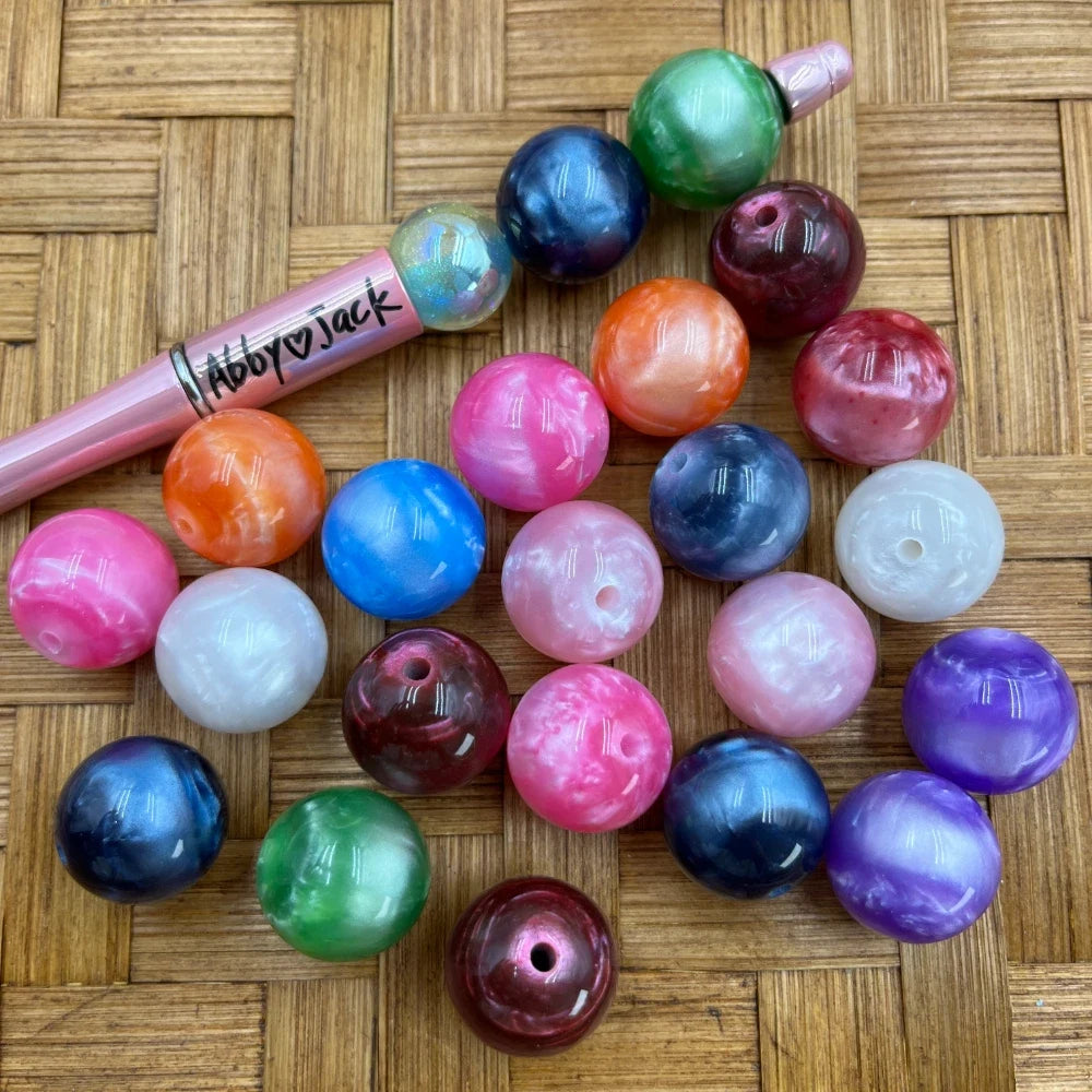 20MM galaxy marble acrylic beads in assorted colors including pink, navy, orange, green, purple, white, and blue with pearlescent swirl finish, displayed on woven surface next to a beadable pen.