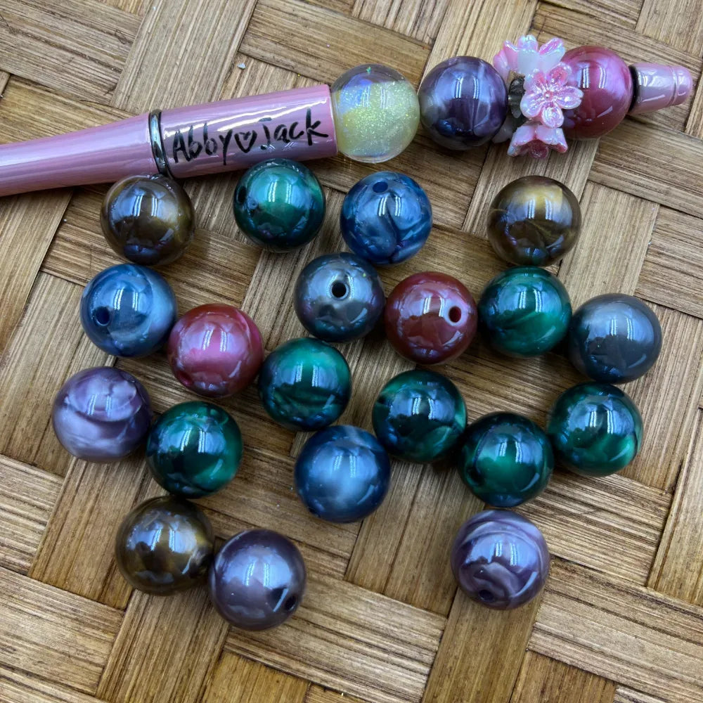 Assorted galaxy marble iridescent acrylic beads in teal, purple, blue, green, and red tones displayed on woven surface next to a pink beadable pen labeled Abby Jack