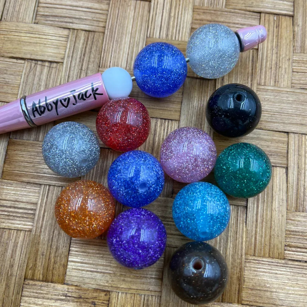 20MM glitter acrylic beads in 12 colors including blue, red, purple, teal, orange, pink, silver, black, and clear displayed on a woven surface next to a pink beadable pen from Abby Jack.