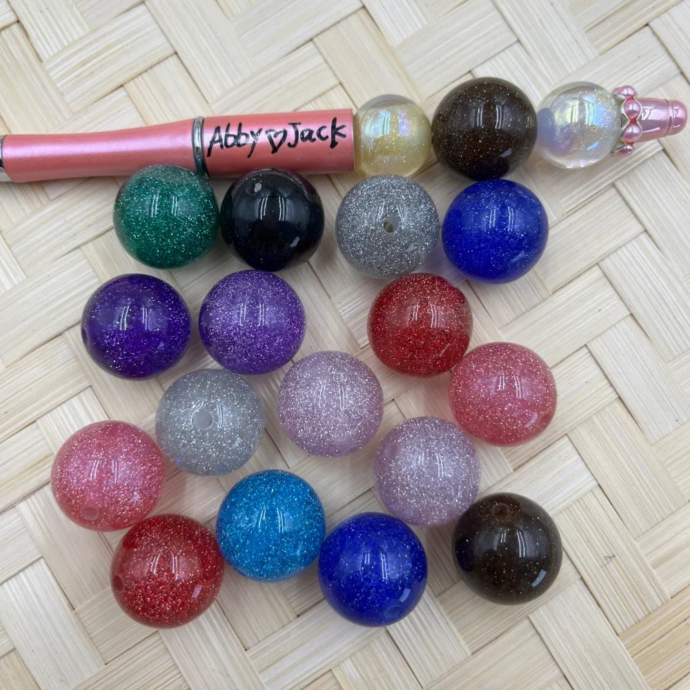 20MM round glitter acrylic beads in assorted colors including green, black, silver, blue, purple, red, pink, and brown displayed on a woven surface next to a beadable pen.