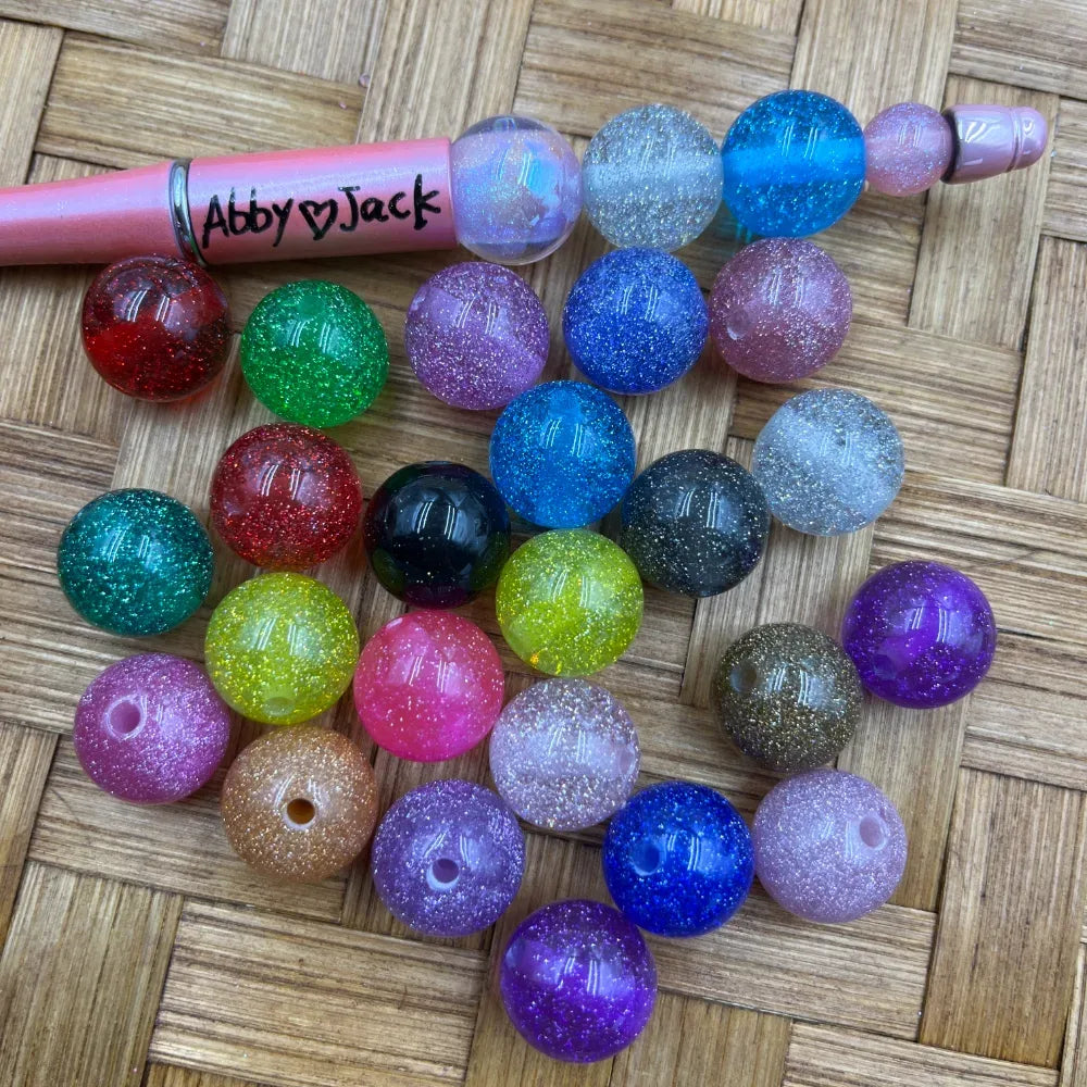Assorted 20mm glitter acrylic round beads in multiple colors including red, green, blue, purple, pink, teal, yellow, black, and clear with sparkle finish, shown next to a pink beadable pen on a woven surface.