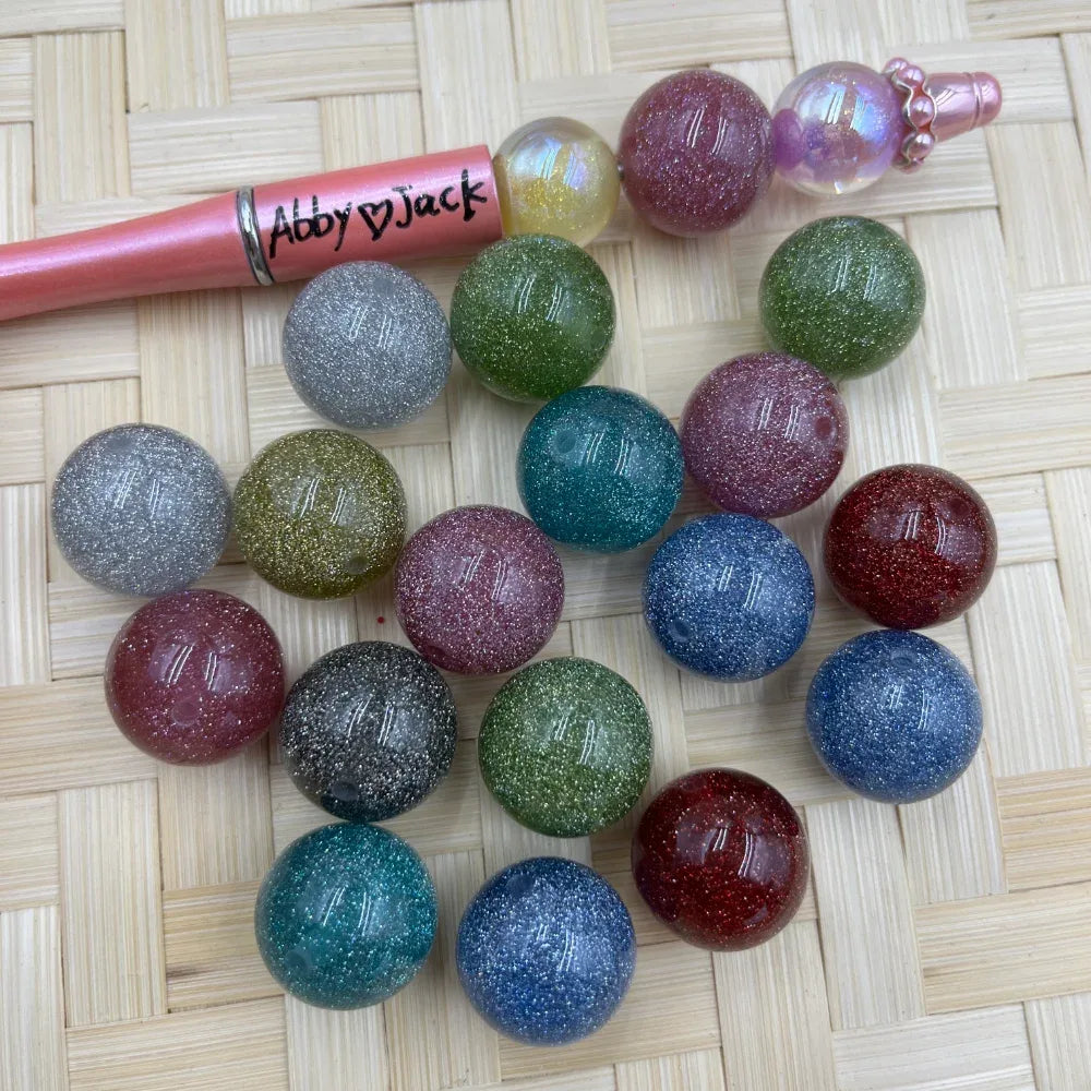 20MM multicolor glitter galaxy acrylic beads in assorted colors including teal, red, blue, green, pink, silver, and gold arranged on a wicker surface next to a beadable pen.