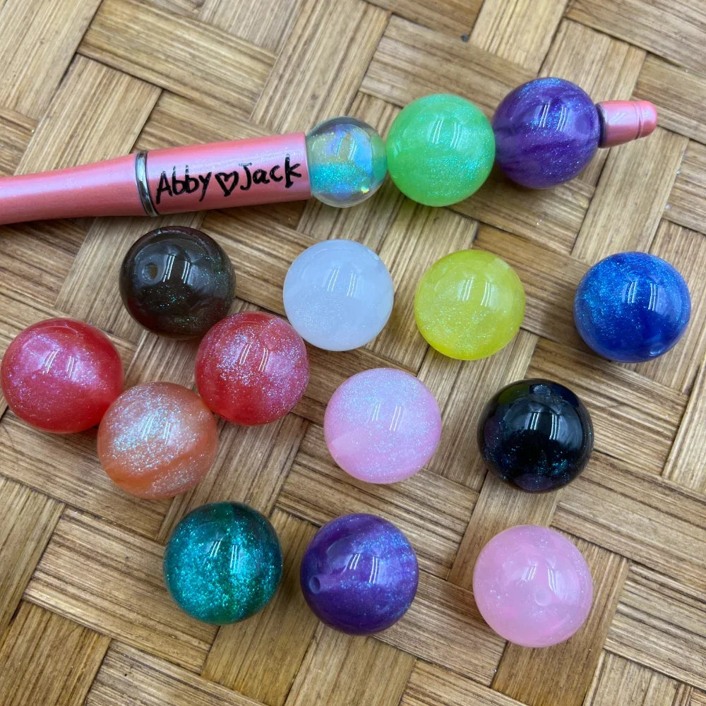 20MM glitter galaxy acrylic beads in assorted colors including green, purple, blue, teal, pink, red, and clear, displayed next to a pink beadable pen on a woven surface.