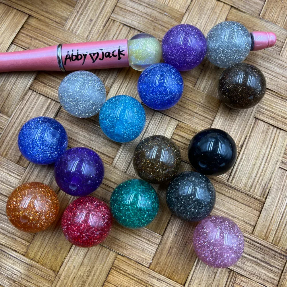 15 assorted 20MM glitter galaxy acrylic beads in multiple colors including blue, purple, teal, red, orange, black, brown, pink, and white, displayed on a woven surface next to a pink beadable pen branded Abby Jack.