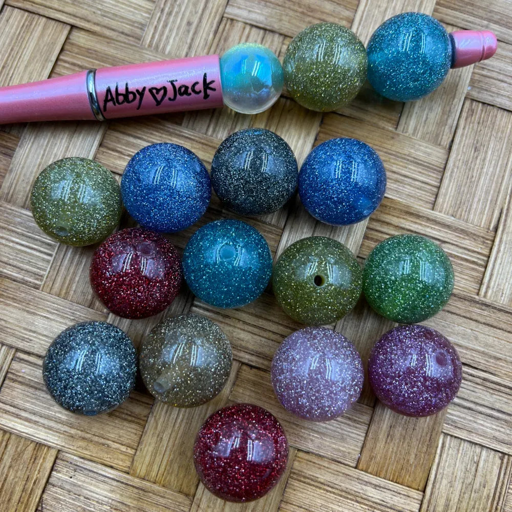 20MM round glitter acrylic beads in assorted colors including blue, green, red, teal, purple, and gray sparkle finish displayed on woven surface next to a beadable pen labeled Abby Jack.