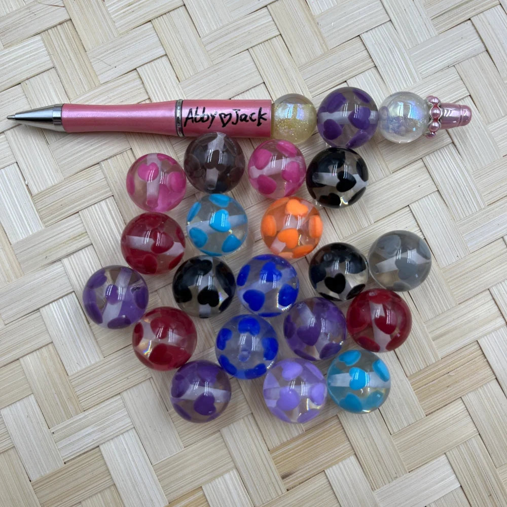 20MM clear acrylic round beads with colorful heart patterns in assorted colors including pink, purple, blue, orange, red and black, displayed next to a pink beadable pen on a woven surface.
