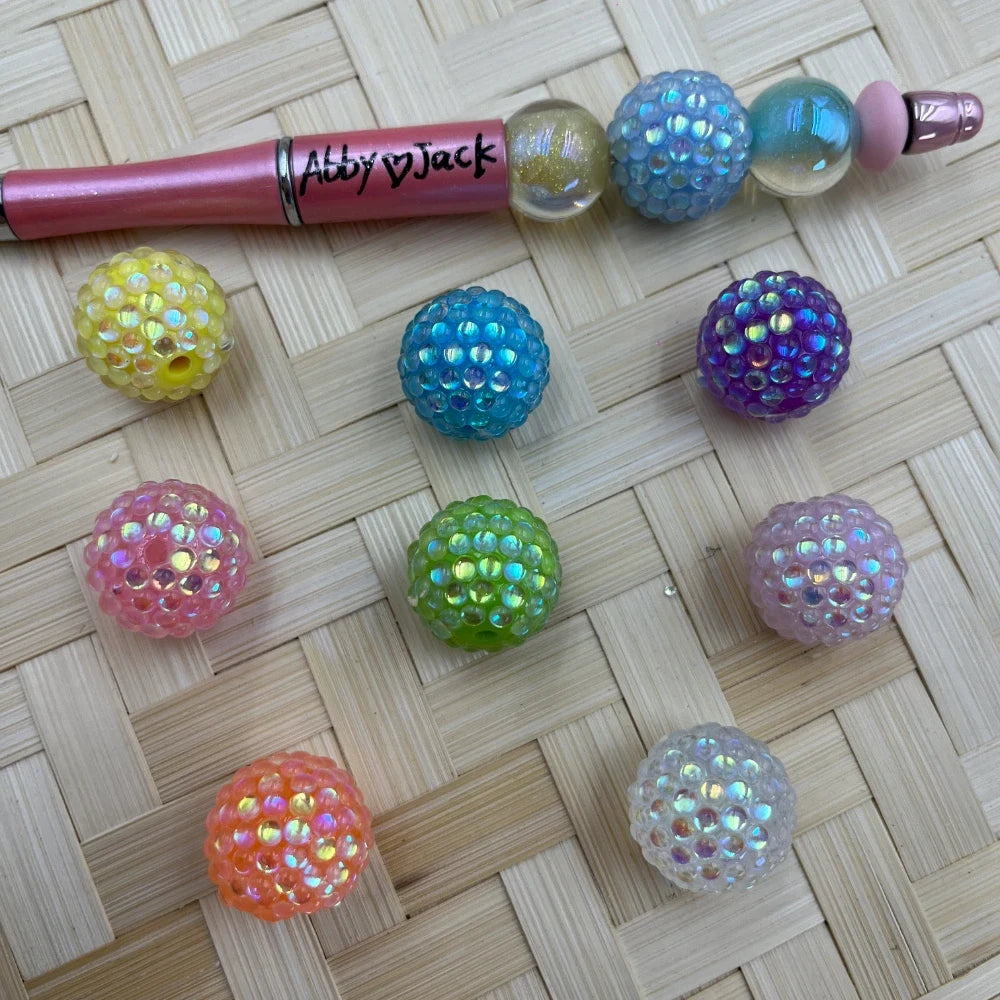Eight iridescent AB bumpy berry acrylic beads in yellow, pink, teal, green, purple, lavender, orange, and white displayed on a woven bamboo surface next to a pink beadable pen.