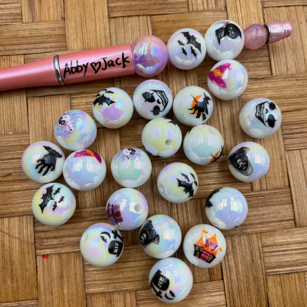Iridescent white acrylic beads with Halloween prints including bats, witches, ghosts, RIP gravestones, and haunted houses, displayed next to a pink beadable pen on a woven surface.