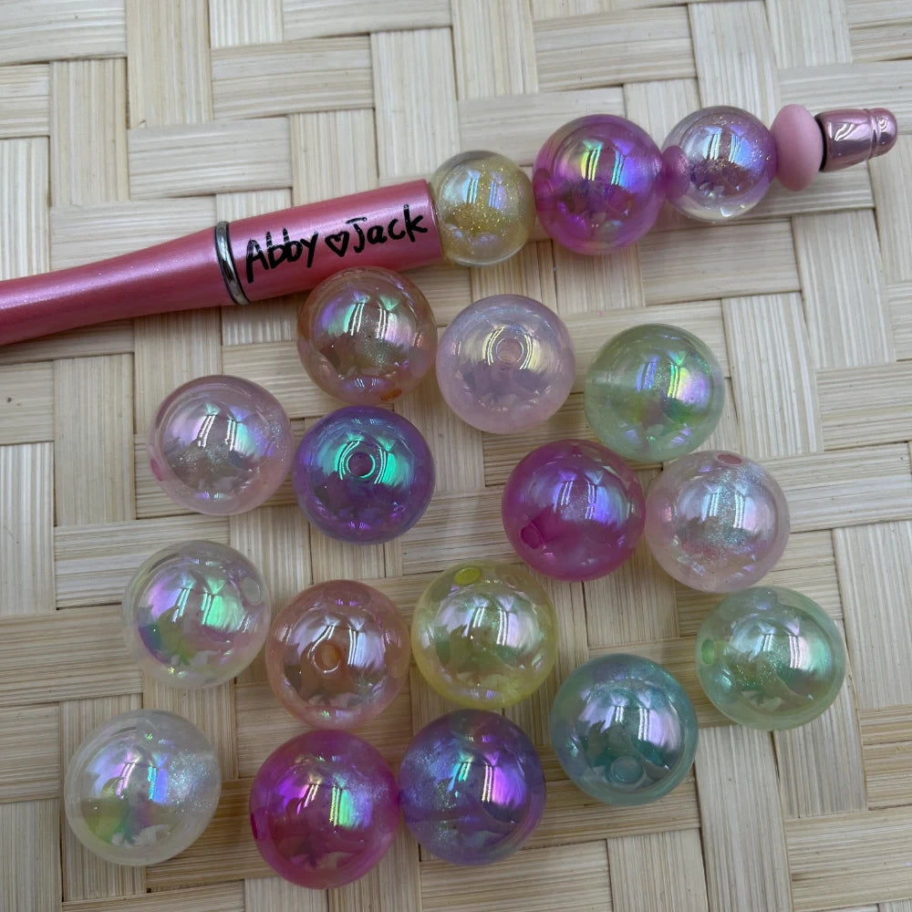 20MM iridescent aurora AB acrylic beads in pastel pink, purple, green, and yellow holographic shimmer finish displayed on woven surface next to a pink beadable pen labeled Abby and Jack.