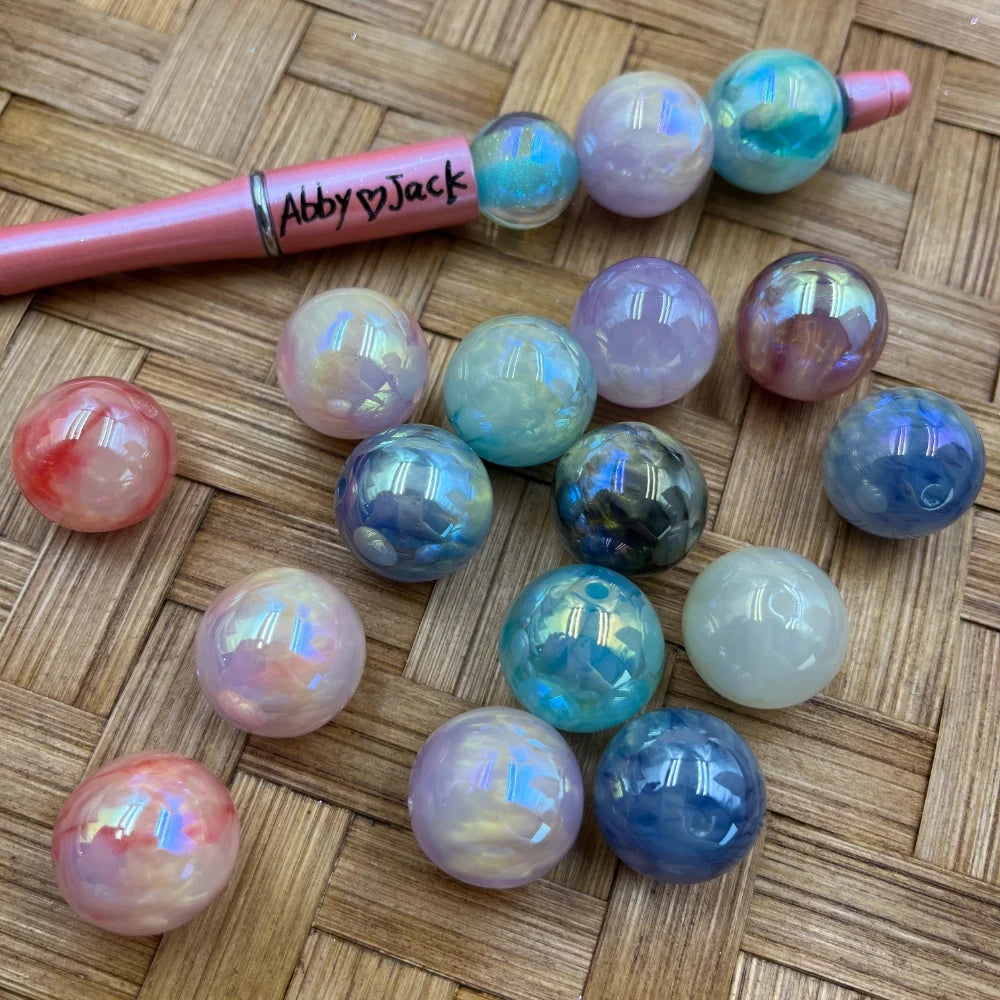 20MM iridescent aurora AB acrylic beads in assorted pastel and jewel tone colors including pink, blue, purple, teal, and white with holographic shimmer finish, displayed on woven surface next to a beadable pen.