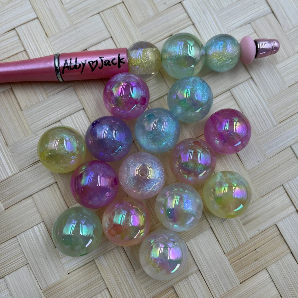 20MM iridescent aurora AB acrylic beads in pastel rainbow holographic colors next to a pink beadable pen on a wicker surface