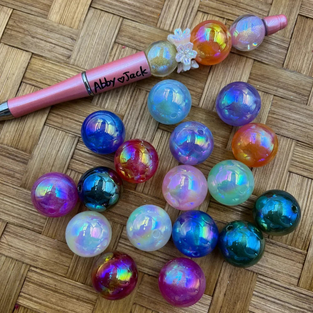 20MM iridescent aurora acrylic beads in assorted colors including purple, blue, pink, orange, teal, white, and red with holographic shimmer finish, displayed alongside a pink beadable pen on a woven surface.