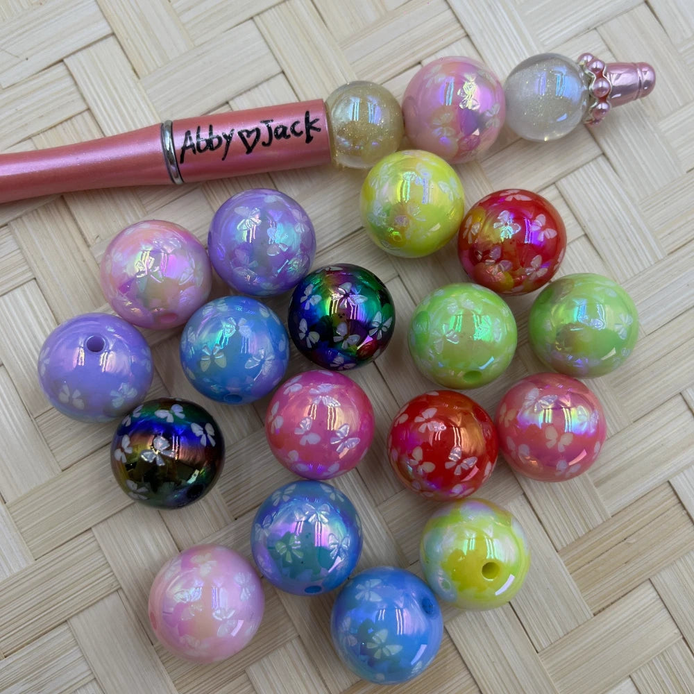 20MM iridescent butterfly printed acrylic beads in assorted colors including purple, pink, red, green, black, and yellow on a woven surface next to a beadable pen.