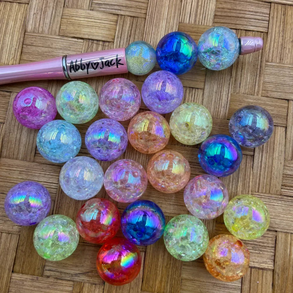 20MM iridescent crackle acrylic beads in assorted colors including blue, purple, pink, red, yellow, and clear with rainbow AB finish, displayed on woven surface next to a pink beadable pen labeled Abby Jack.