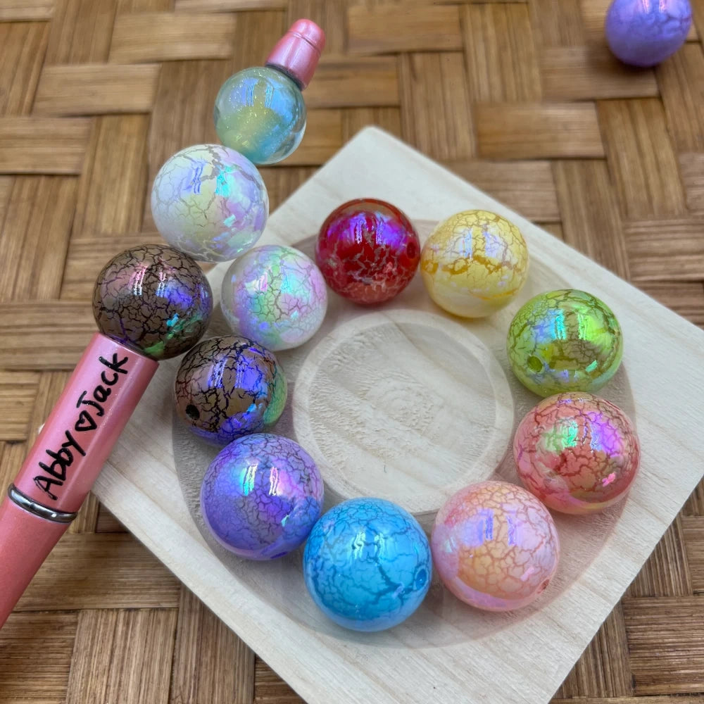 20MM iridescent crackle acrylic beads in assorted colors including blue, purple, pink, red, green, yellow, and brown arranged in a circle on a wood tray next to a pink beadable pen labeled Abby Jack