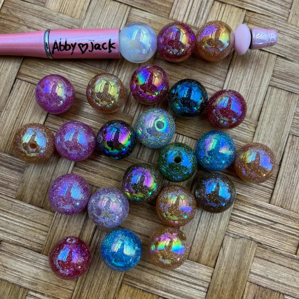 Assorted iridescent galaxy glitter acrylic beads in purple, teal, red, brown, and black with holographic AB finish displayed next to a pink beadable pen on a woven surface