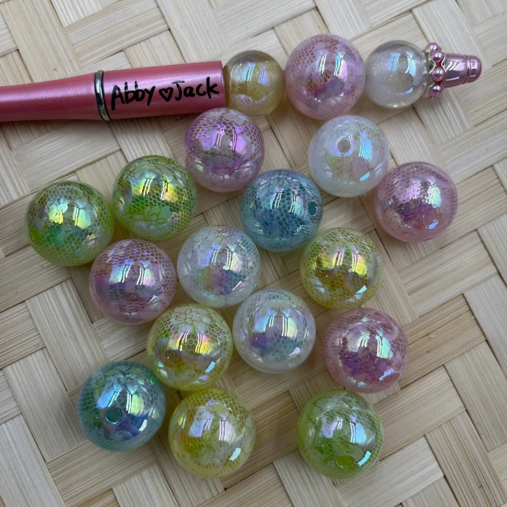 20MM iridescent mermaid scale acrylic beads in pastel pink, green, teal, and white with holographic shimmer finish, shown next to a beadable pen on a wicker surface.