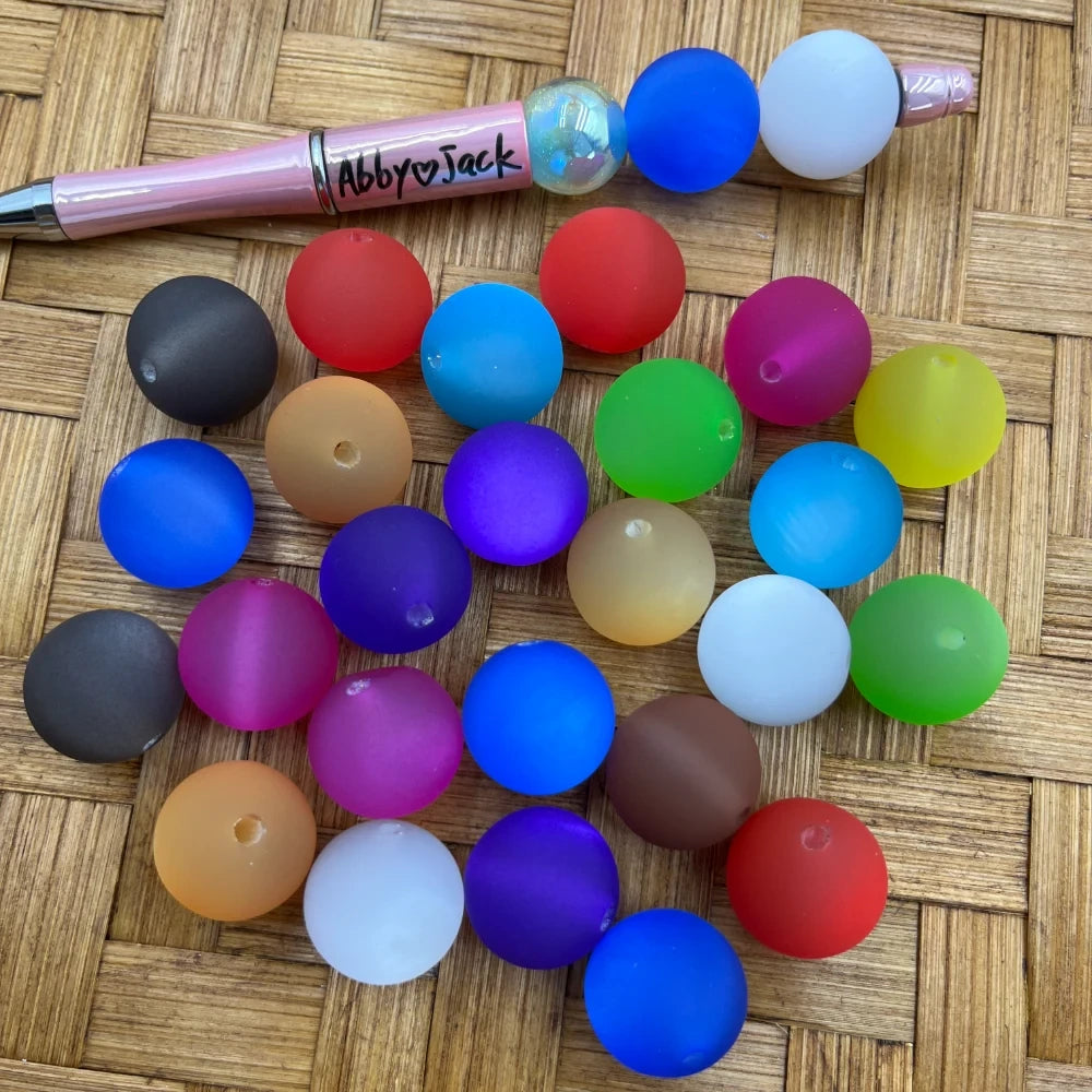 20MM matte frosted round acrylic beads in assorted colors including blue, red, purple, green, yellow, white, black, orange, and pink displayed on a wicker surface next to a pink beadable pen.