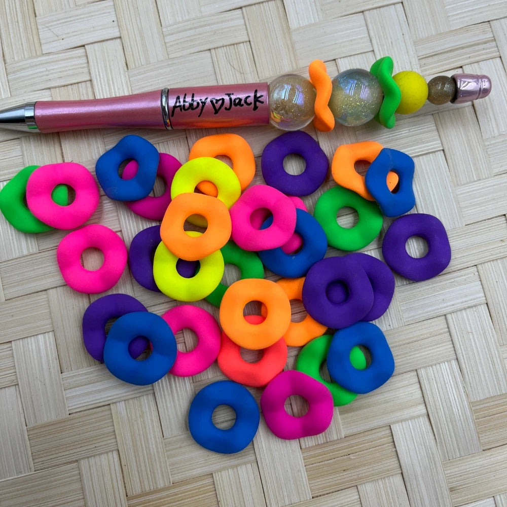 20MM neon donut ring acrylic spacer beads in pink, orange, yellow, green, blue, and purple scattered on a woven surface next to a beadable pen.
