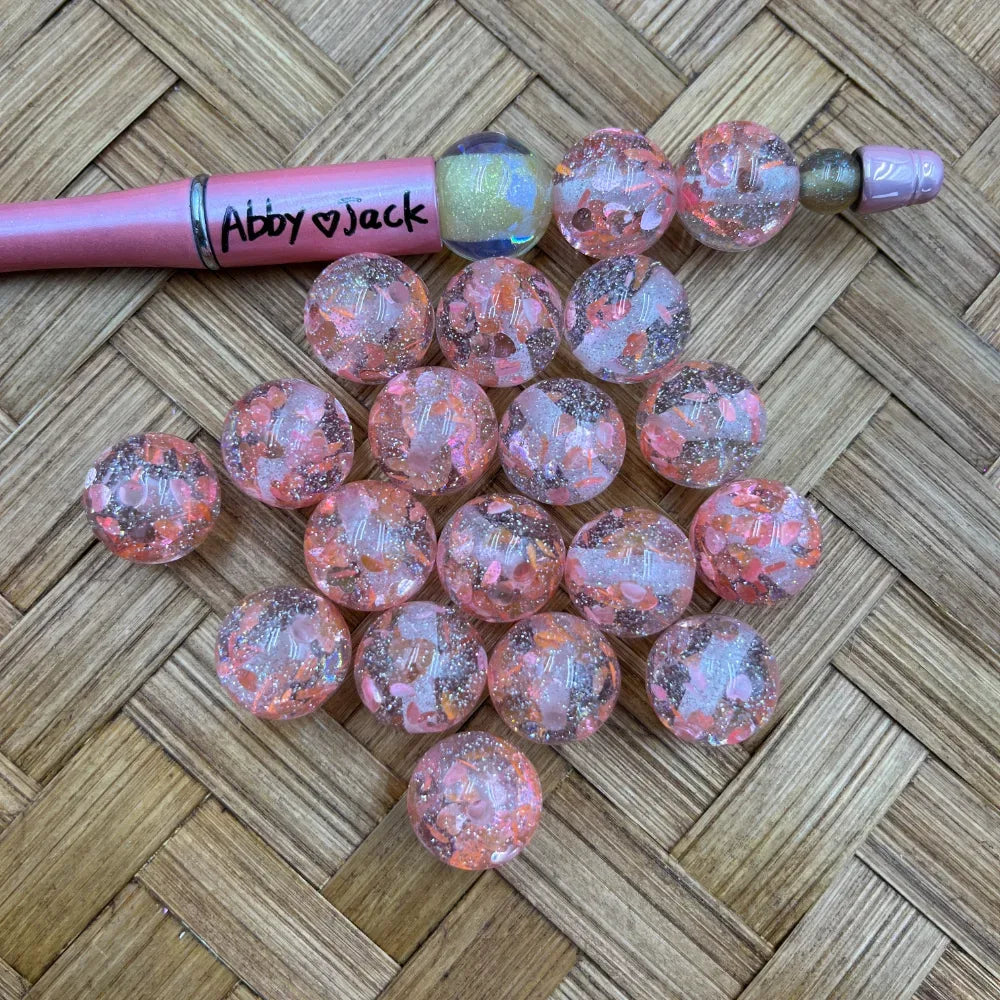 Chunky round pink glitter confetti acrylic beads with rose gold foil flakes displayed on wicker surface next to a pink beadable pen labeled Abby Jack