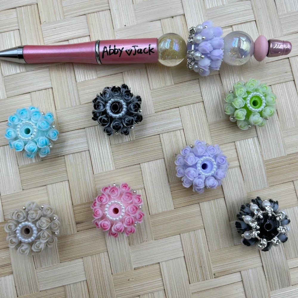 Assorted rose wreath resin fancy beads in pink, blue, lavender, green, white, and black with pearl and rhinestone accents beside a decorated beadable pen.