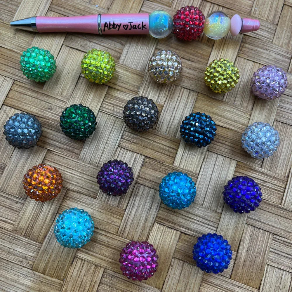20MM assorted color rhinestone disco ball beads displayed on a woven surface next to a pink beadable pen, available in green, teal, purple, orange, blue, pink, yellow, gray, and more.