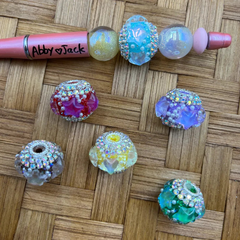 Assorted rhinestone-trimmed galaxy glitter resin fancy beads in red, purple, yellow, teal, clear, and iridescent colors with floral star details, displayed on a woven rattan surface next to an Abby Jack beadable pen for size reference.